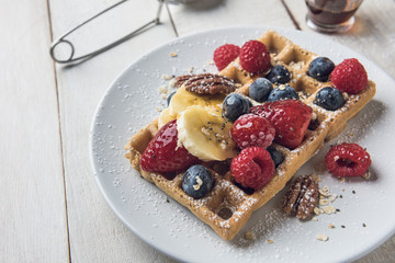 Homemade waffle with banana slices, oat, pecans and berries served on a white plate over a white...