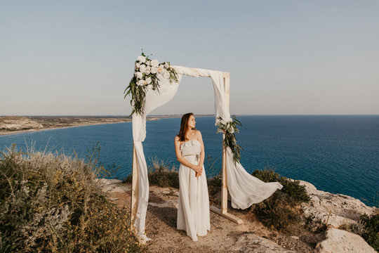 bride in a wedding dress stands in front of a wedding arch on a cliff above the sea