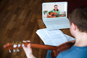 Man playing acoustic guitar and watching online lesson on laptop while practicing at home. Stay...