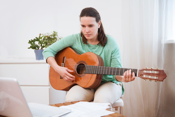 Woman playing acoustic guitar and watching online lesson on laptop while practicing at home. Stay home. quarantine. Online training, online classes.
