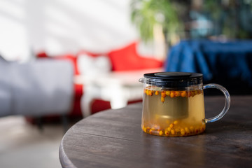 Tasty tea with sea buckthorn on a table in a decorated room. Hot berry drink in a glass teapot.