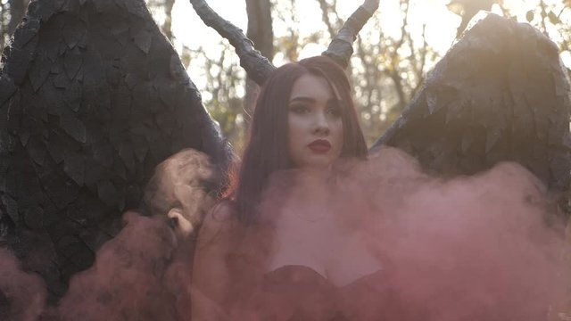 Pretty sexy awesome brunette girl maleficent witch walking in the forest among trees with red smoke having horns and wings in the sunset