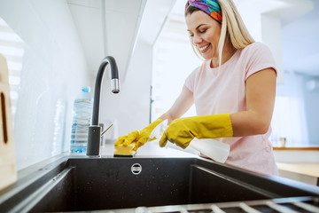 Worthy smiling dedicated housewife with rubber gloves on spraying and cleaning with sponge kitchen...