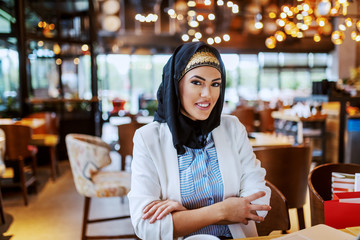 Charming modern smiling Muslim woman with scarf sitting in cafeteria with hands folded and looking at camera.
