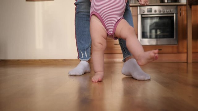 Mother supports toddler baby child makes first steps barefoot on the floor at home