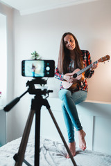 Girl in a shirt and jeans writes a vlog on the phone about playing ukulele.