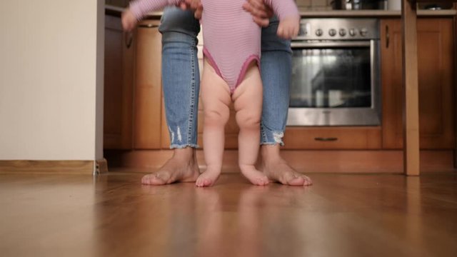 Mother supports play with toddler baby child makes first steps barefoot on the floor at home