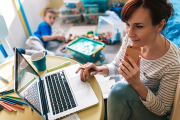 Mother working remotely on laptop while taking care of her son playing with toys in his room.