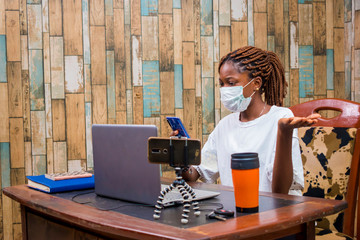 young black woman sitting at home, wearing a nose mask, vlogging with her smartphone while working on her laptop