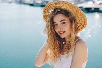 curly blonde with a tender face in a straw hat smiles against the backdrop of a sparkling sea, boats are blurry away in the distance