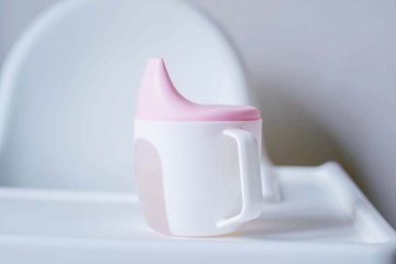 Baby cup with lid. White cup with pink lid. Tableware for infants and children in the first year of life. Thirst, drinks, dehydration.