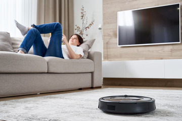Robotic vacuum cleaner cleaning the room while woman resting on sofa