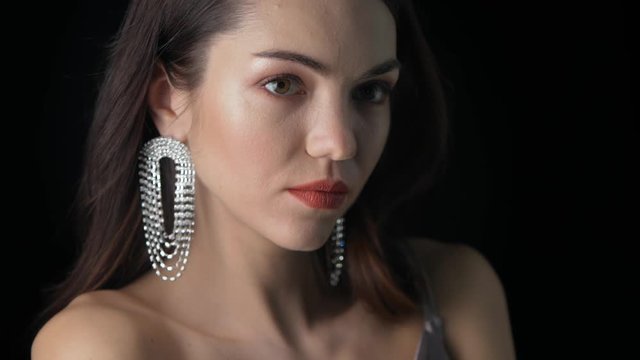 Young beautiful model poses for the photographer at a photo shoot in the studio on a black background, close-up. Brunette with makeup and shiny gemstone earrings under the camera flashes.