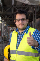 Positive and successful young construction worker posing showing thumbs up gesture. Outdoors