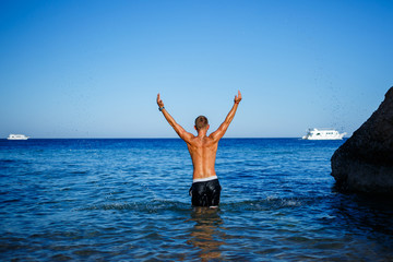 Obraz premium Happy man guy joy on vacation, stands at sea and splashes in the water. Men's shorts, sun, beach