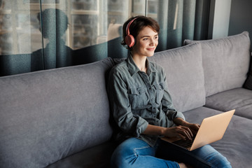 Image of nice young smiling woman using laptop and headphones - obrazy, fototapety, plakaty