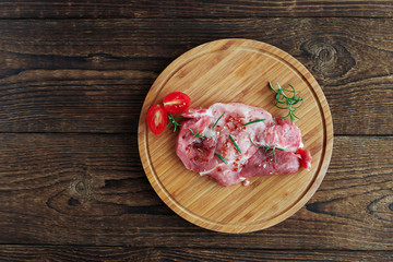 On a round wooden cutting board lies a raw pickled pork steak. Near a sprig of rosemary and cherry...