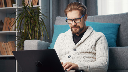 Middle-aged male working from home sitting on floor leaning against couch using laptop, quarantine