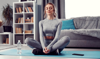 Meditative lady with closed eyes sitting with folded legs on exercise mat on floor listening to...