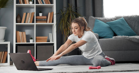 Young woman sitting on floor watching online lesson on laptop and doing stretching in apartment
