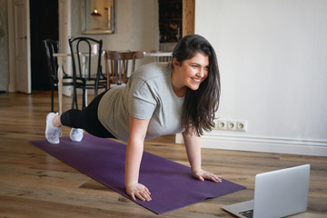 Sporty plus size woman in sportswear working out at home, doing plank on yoga mat in front of open...