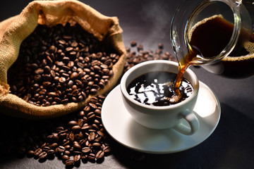 Pouring coffee with smoke on a cup and coffee beans on burlap sack on black background.