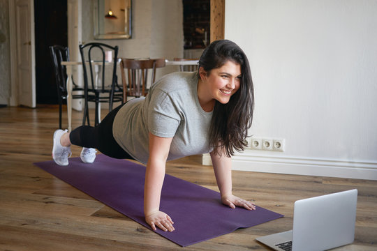 Sporty plus size woman in sportswear working out at home, doing plank on yoga mat in front of open laptop, repeating instructions by professional fitness trainer watching online video tutorial