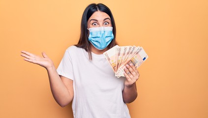 Young woman wearing protection mask for coronavirus holding 50 euros banknotes celebrating achievement with happy smile and winner expression with raised hand