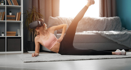 Slim girl in VR and activewear side-lying on floor raising one leg up doing fitness exercises in apt - obrazy, fototapety, plakaty