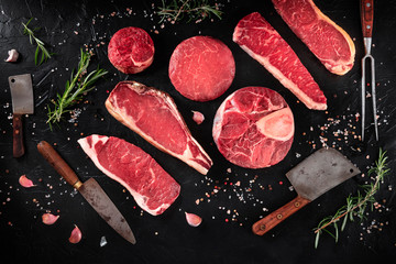 Cuts of meat, shot from the top on a black background with salt, pepper, rosemary and knives, a flat lay - obrazy, fototapety, plakaty