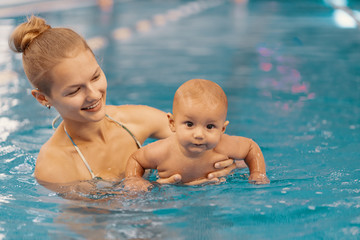 Fototapeta na wymiar Young mother and her baby enjoying a baby swimming lesson in the pool. Child having fun in water with mom