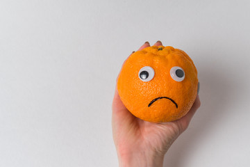 Female hand holding sad orange. Funny orange fruit character. Mandarin-smile