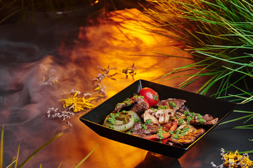 Grilled vegetables: mushrooms, bell pepper, eggplant with grilled meat  in a black square matte plate among greens on a gray smoky background with warm lighting. Space. Close up