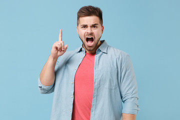 Angry irritated young bearded guy 20s in casual shirt posing isolated on pastel blue background in studio. People lifestyle concept. Mock up copy space. Screaming swearing pointing index finger up.