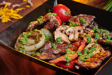 Grilled vegetables: mushrooms, bell pepper, eggplant with grilled meat  in a black square matte plate among greens on a gray smoky background with warm lighting. Space. Close up