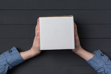 Children's hands holds white square cardboard box. Black background. Top view. Shipping concept.