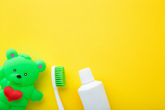 Green rubber teddy bear with red heart, toothbrush and tube of toothpaste on bright yellow table background. Children teeth cleaning. Closeup. Empty place for text.