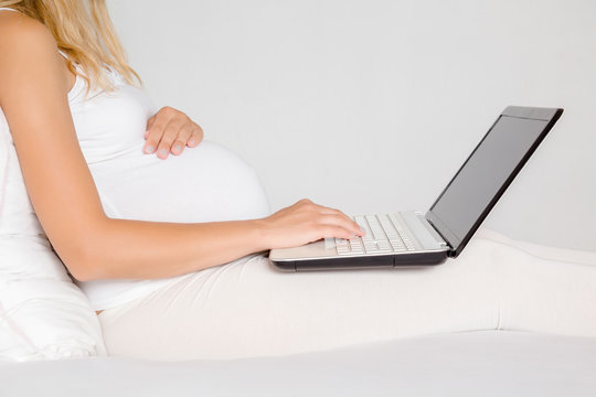 Pregnant woman in white clothes sitting and using laptop in bed. Baby expectation. Homely atmosphere. Pregnancy information searching concept. Close up. Side view.