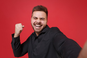 Obraz premium Close up of happy young bearded guy 20s in classic black shirt posing isolated on red background. People lifestyle concept. Mock up copy space. Doing selfie shot on mobile phone, doing winner gesture.