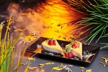 Delicious Cheesecake, decorated with strawberries on a square black plate in a dark smoky background with warm lighting. Beautiful greens on the background. Close up. Space