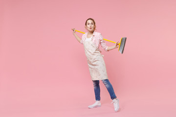 Amazed pretty young woman housewife in casual clothes, apron doing housework isolated on pastel pink wall background studio portrait. Housekeeping concept. Mock up copy space. Hold in hands broom.