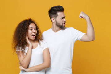 Excited young couple two friends european guy african american girl in white t-shirts posing isolated on yellow background studio. People lifestyle concept. Mock up copy space. Showing biceps muscles.