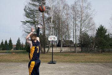 Photo of young basketball player practicing in the sport ground with trees on background. The ball flies into a basketball hoop. Back view