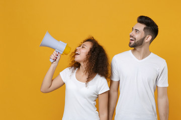 Cheerful couple two friends european guy african american girl in white t-shirts posing isolated on yellow background. People lifestyle concept. Mock up copy space. Scream in megaphone, looking aside.