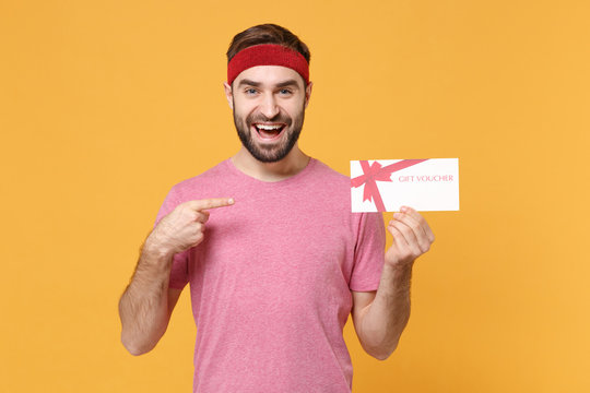 Funny young bearded fitness sporty guy sportsman in headband t-shirt in home gym isolated on yellow background. Workout sport motivation lifestyle concept. Pointing index finger on gift certificate.