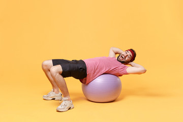 Fototapeta na wymiar Excited young bearded fitness sporty guy 20s sportsman in headband t-shirt in home gym isolated on yellow background. Workout sport motivation lifestyle concept. Doing abdominal exercises on fitball.