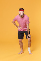 Ingured displeased young bearded fitness guy sportsman in headband t-shirt in home gym isolated on yellow background. Workout sport motivation concept. Touching leg with elastic bandage on ankle foot.