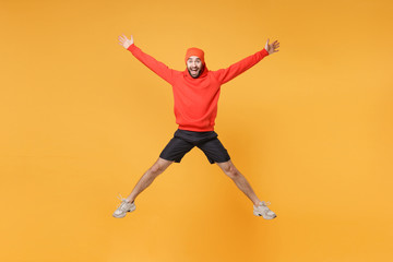 Fototapeta na wymiar Excited young bearded fitness sporty guy 20s sportsman in hat hoodie shorts in home gym isolated on yellow background. Workout sport motivation lifestyle concept. Jumping spreading hands and legs.