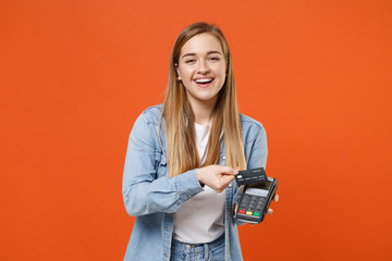 Cheerful young woman in denim clothes isolated on orange background. People lifestyle concept. Mock up copy space. Hold wireless modern bank payment terminal to process acquire credit card payments.