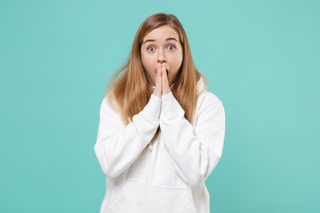 Shocked young woman girl in casual white hoodie posing isolated on blue turquoise background studio portrait. People sincere emotions lifestyle concept. Mock up copy space. Covering mouth with hands.